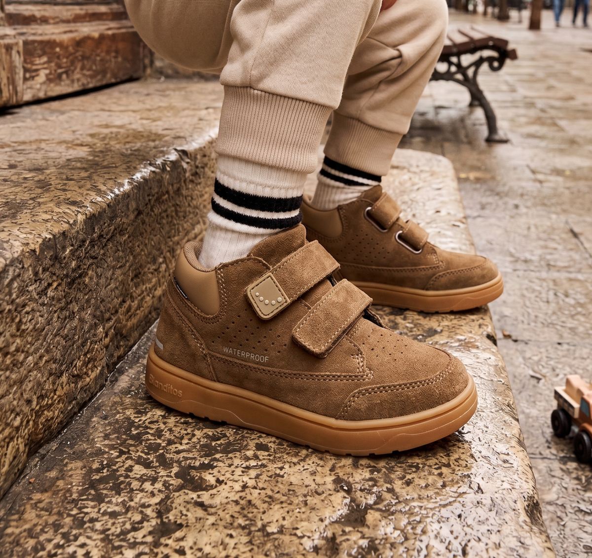 A child wearing tan suede-style sneakers with double hook-and-loop straps, beige pants, and striped socks, sitting on stone steps outdoors.